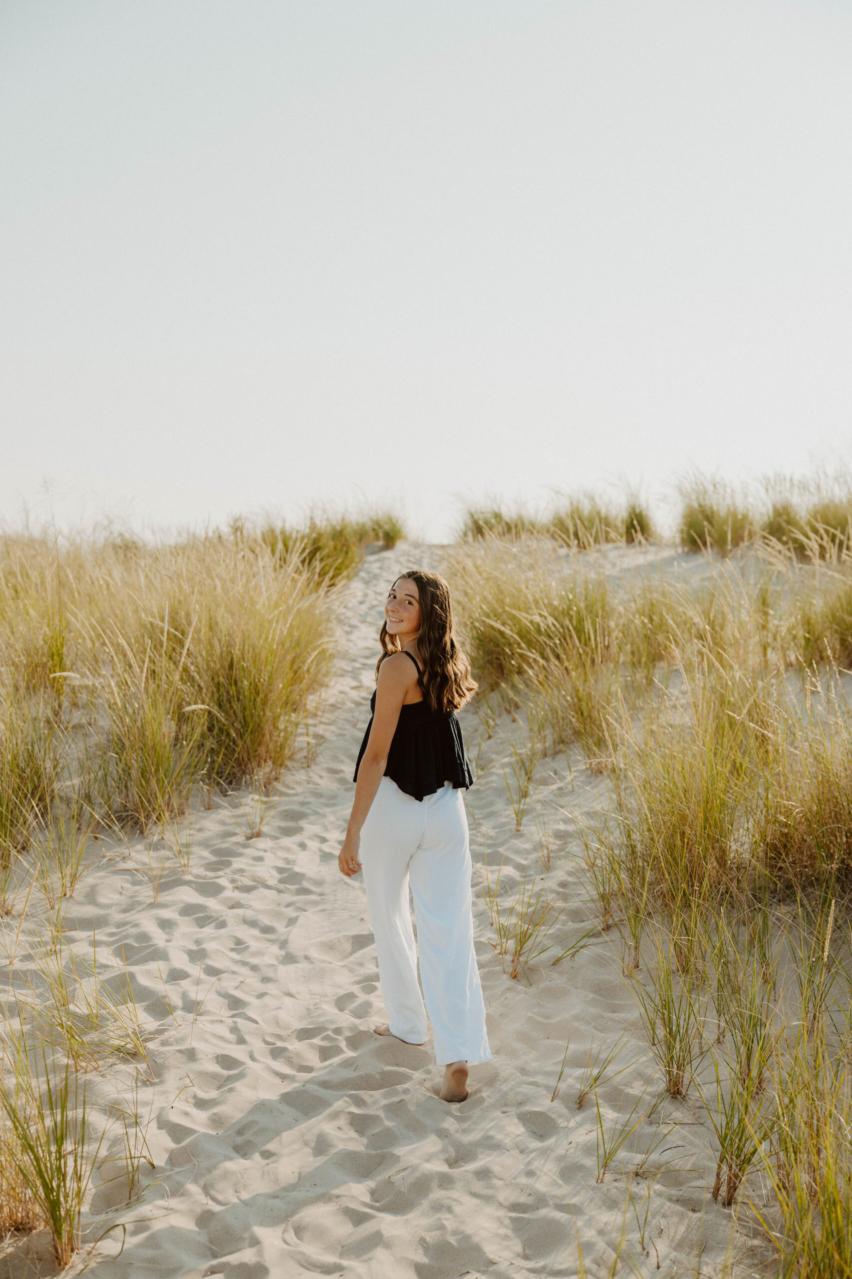 Senior walking the dunes at Ludington beach in white pants and blue tank top, surrounded by dune grass
